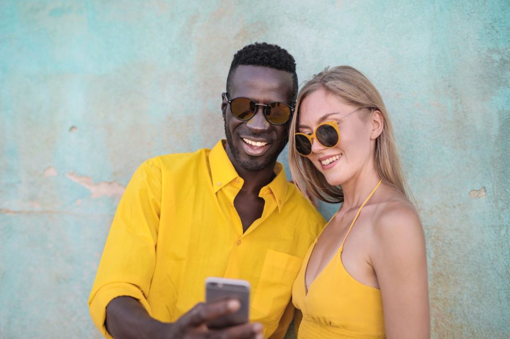 Smiling couple in bright yellow outfits taking a selfie against a textured turquoise wall.